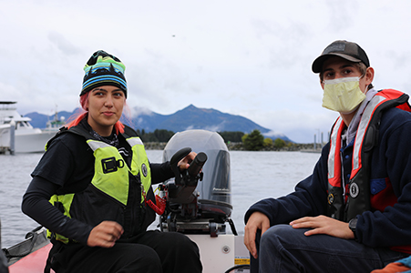 A student, left, operates an outboard engine with a mountainous background in Crescent Harbor with a skiff instructor, right.